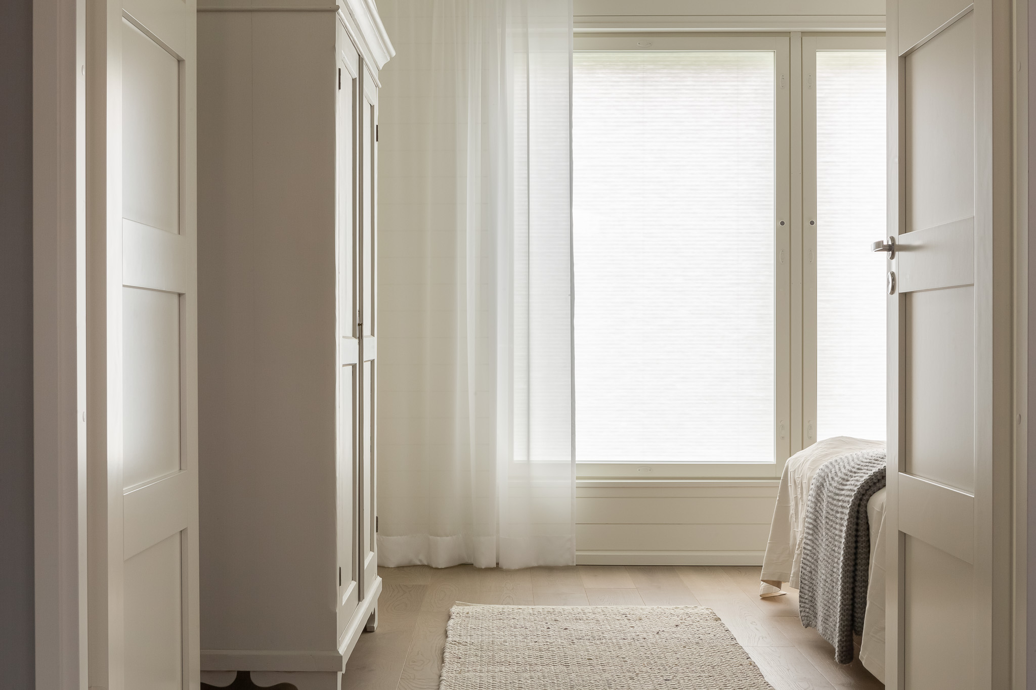 Minimalist bedroom interior with light wooden flooring, a bed, and wide windows fitted with light-filtering pleated blinds.