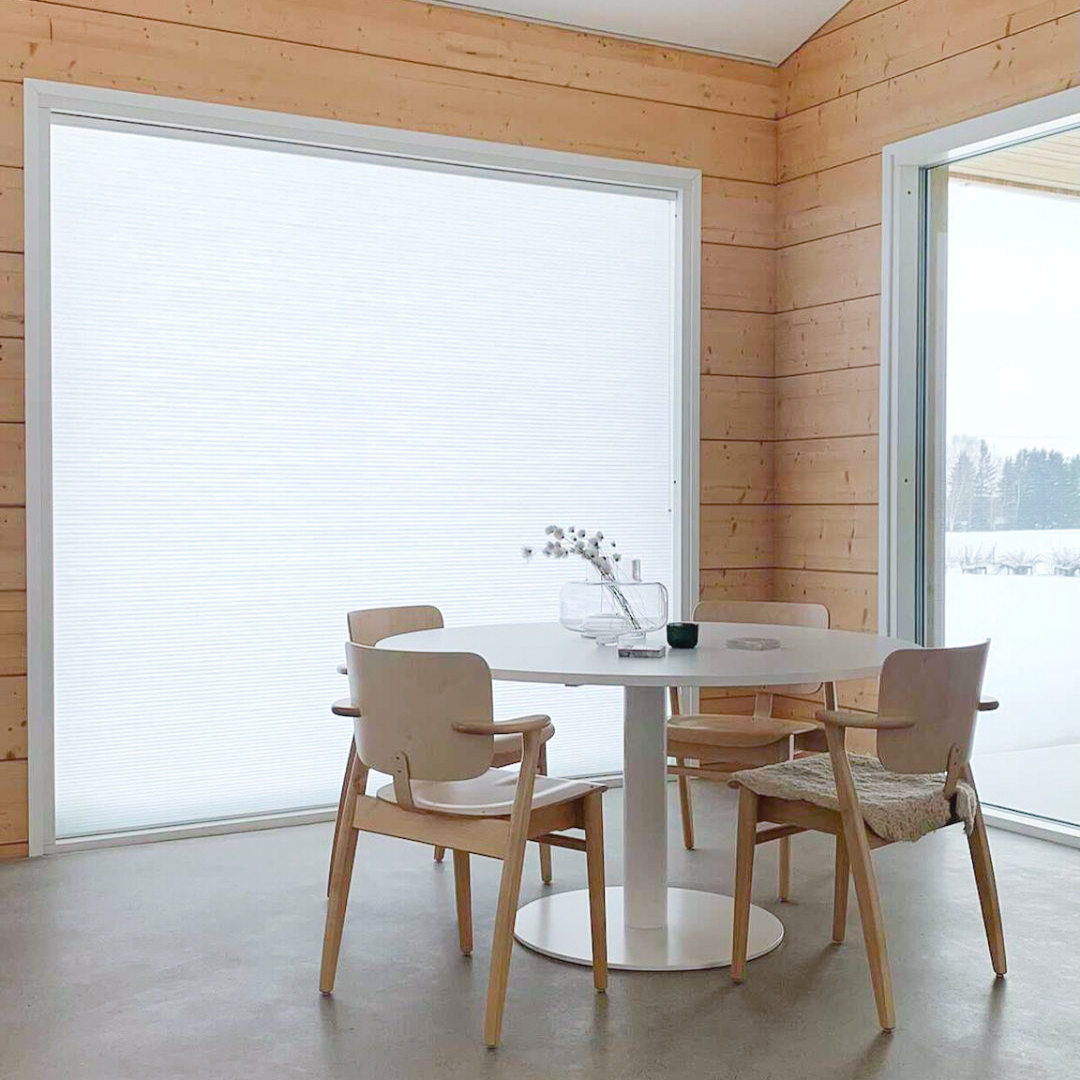 Scandinavian-style dining space featuring light wood interior, simple chairs, and wide windows with light-filtering pleated blinds.