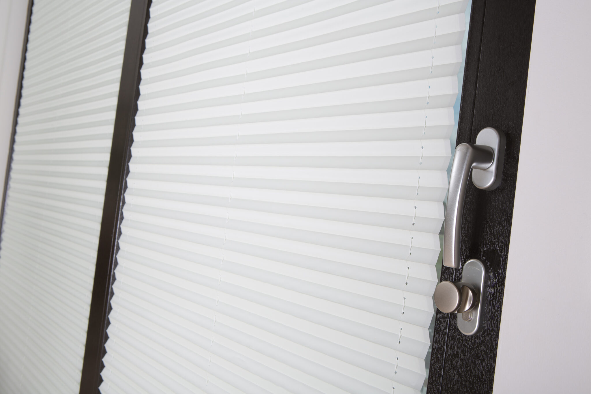 Detail of a window with a light pleated blind fully closed, showing a sleek handle and dark wood frame.