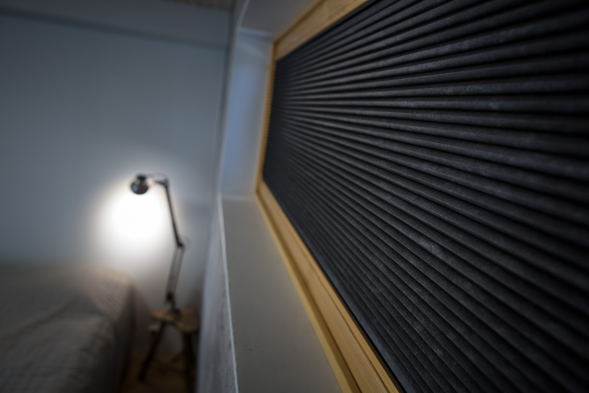 Close-up of dark pleated blind fabric in a window, with a softly lit bedroom and reading lamp in the background