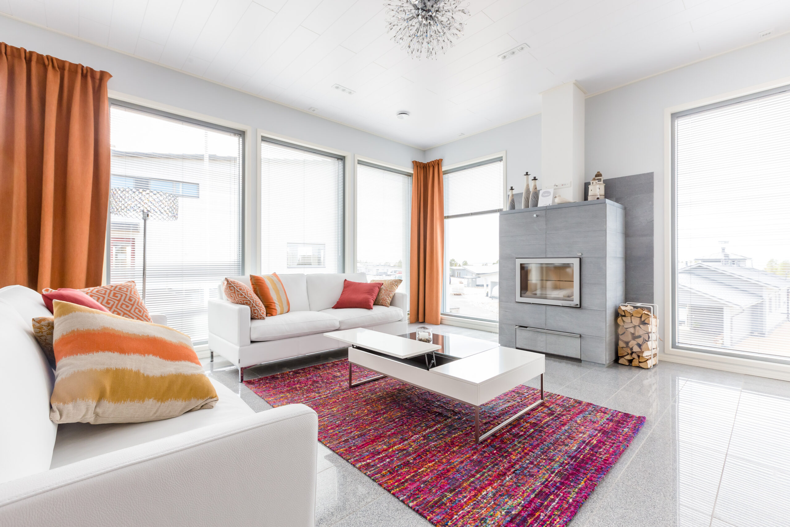 Modern living room with white sofas, colorful rug, and large windows fitted with white venetian blinds, allowing soft natural light into the space