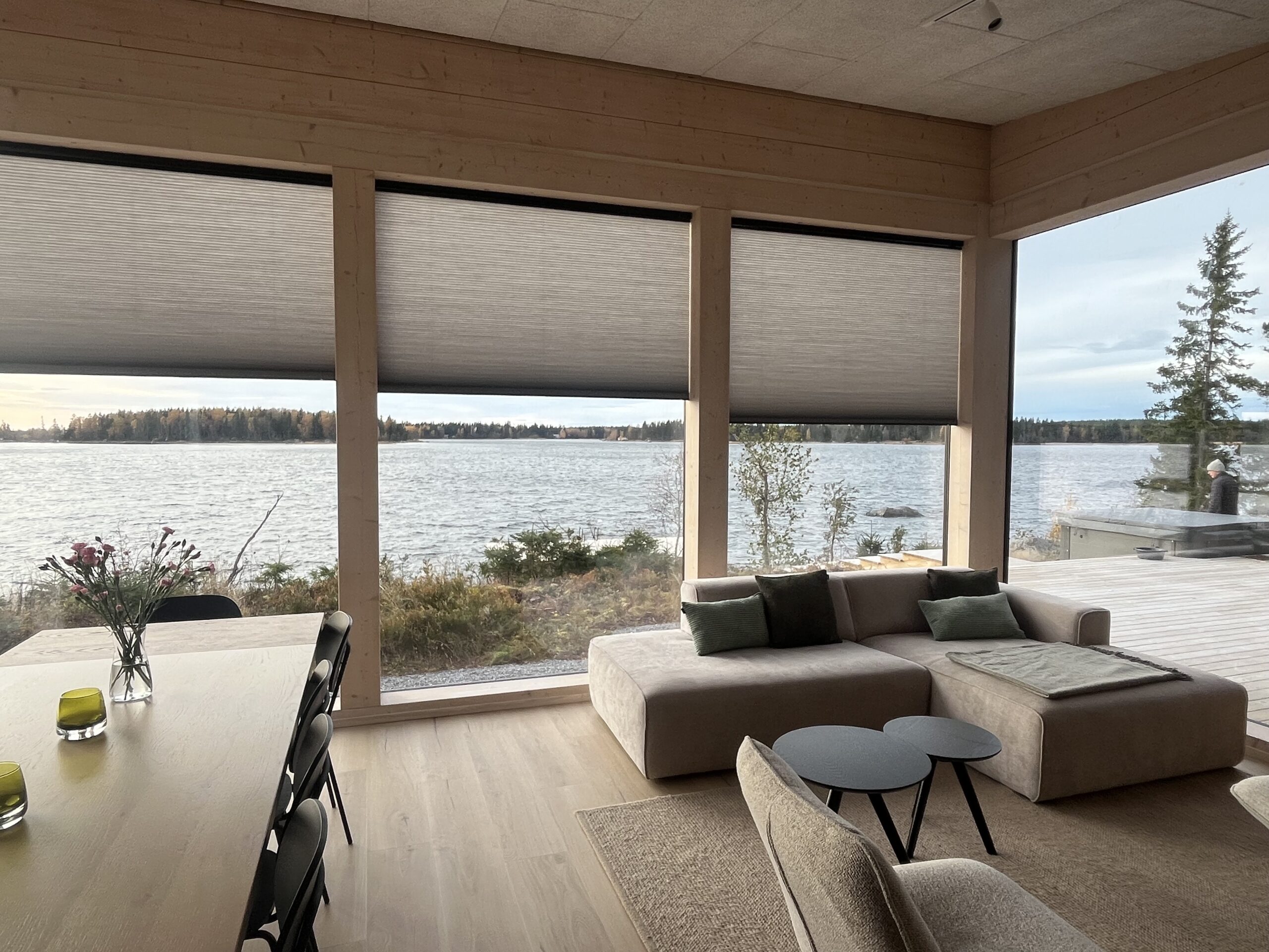 Bright living space with floor-to-ceiling windows, beige sofa and dining area, featuring partially closed pleated blinds and a scenic lake view outside