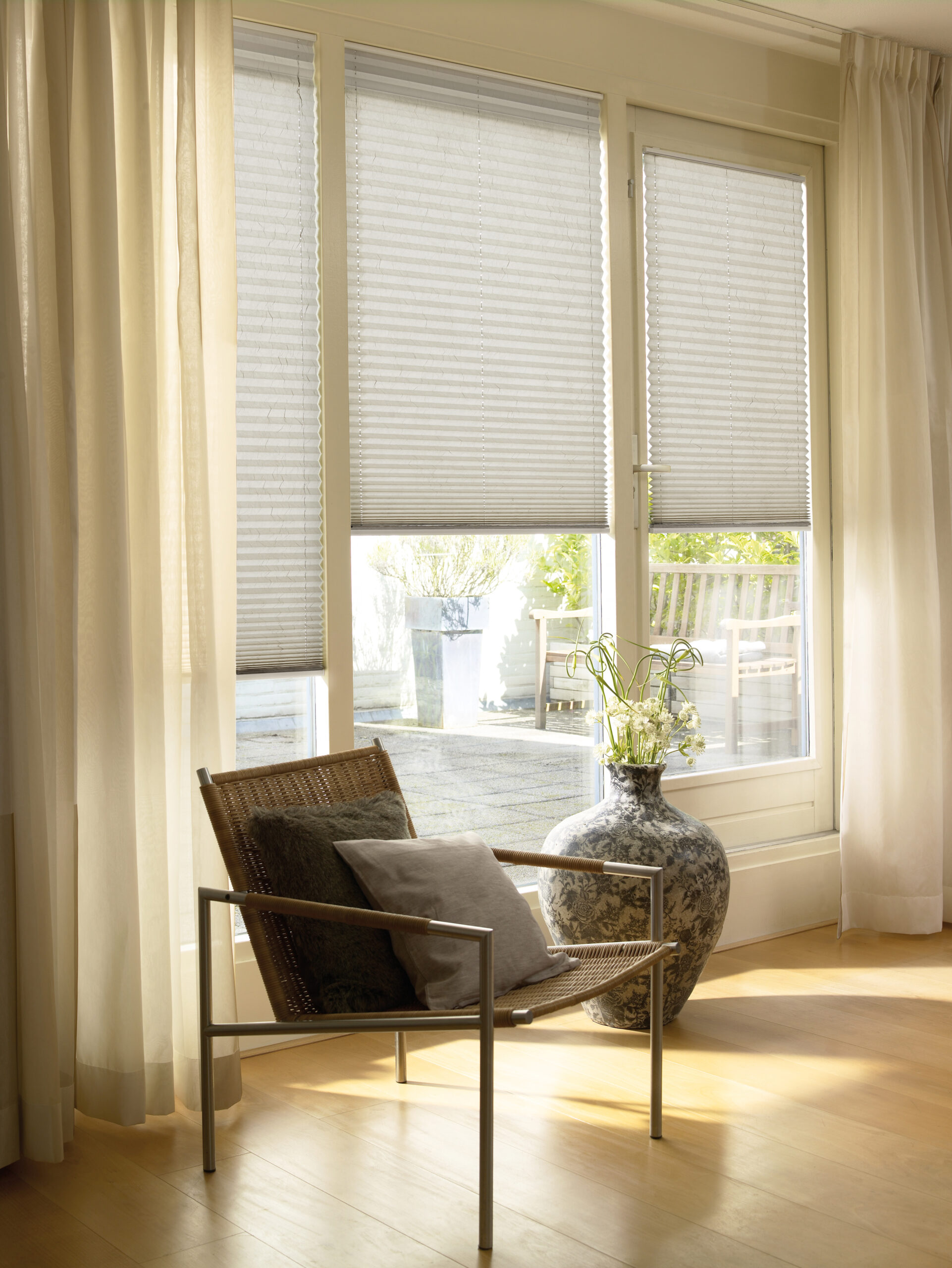 Sunlit living room corner with a chair and vase by large windows fitted with pleated blinds, creating a warm and airy atmosphere
