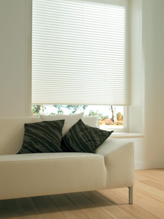 Light-colored pleated blinds partially lowered on a window above a white sofa with dark cushions, gently diffusing natural daylight into the room