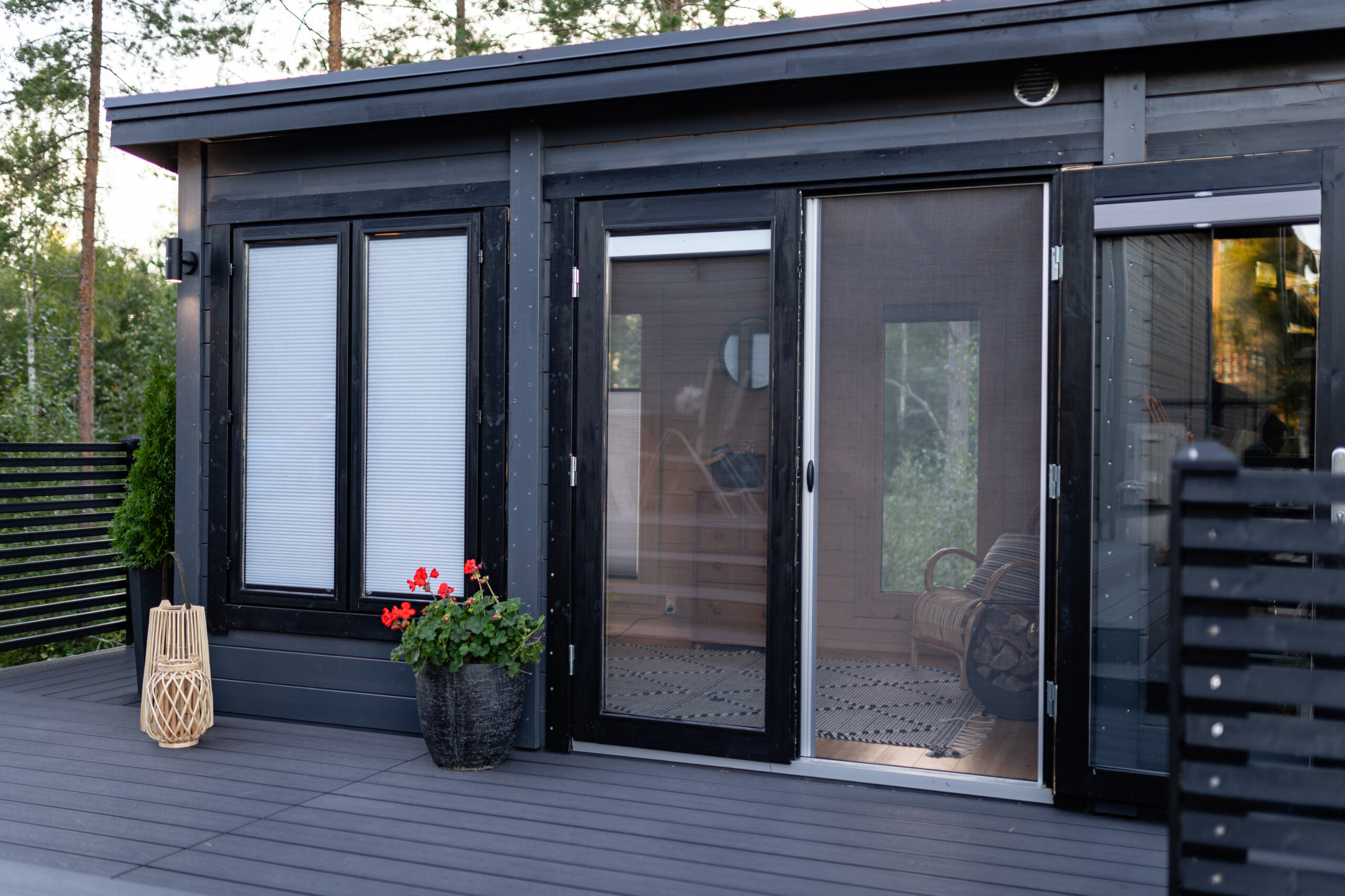 Ett modernt hus med mörk fasad och stora glasdörrar, med plisségardiner på fönstren och en dörr med insektsnät som leder ut till en terrass