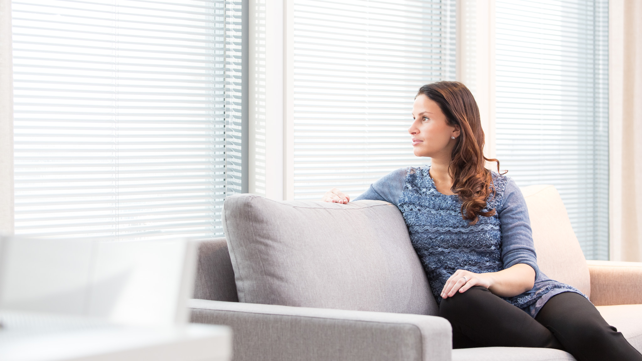 Woman sitting on a sofa by large windows with venetian blinds, enjoying soft natural light in a calm interior